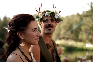 A couple share an intimate moment outdoors with one wearing an antlered floral crown against a forest backdrop.