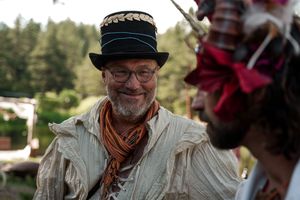 A festival attendee wearing a top hat and scarf smiles at an outdoor medieval gathering.