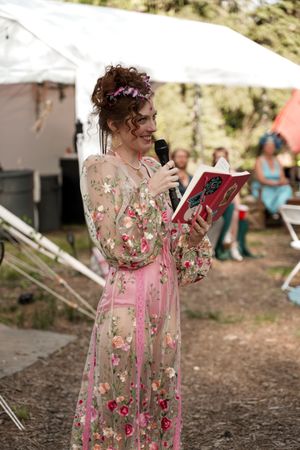 A woman in a floral pink dress reads from a book at an outdoor festival with tents in the background.