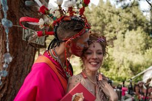 Two people in colorful festival attire share a joyful moment under a tree decorated with red flowers.