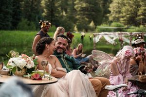 Wedding guests gather at rustic outdoor reception table surrounded by forest scenery with floral centerpieces and place settings.