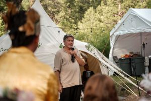The outdoor marketplace features rows of white vendor tents and patrons browsing under a canopy of green trees.