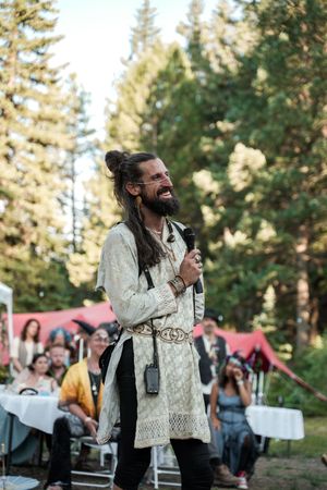 A performer in an ornate tunic laughs joyfully while entertaining guests at an outdoor forest celebration.