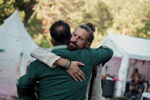 Two men in green shirts embrace warmly at an outdoor event with white tents in the background.