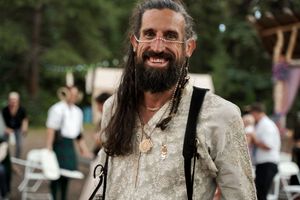 A bearded man in vintage-style attire smiles at an outdoor gathering.