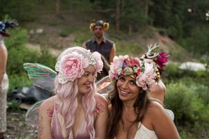 Two friends wearing pink floral crowns and delicate dresses share a joyful moment in a natural outdoor setting.