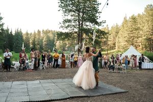 Newlyweds share their first dance on an outdoor dance floor surrounded by wedding guests and pine trees.