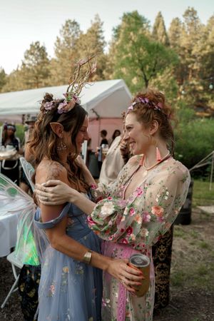Friends wearing floral dresses and flower crowns laugh together at an outdoor celebration.