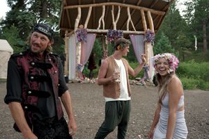 Wedding guests stand near a rustic wooden arch decorated with purple flowers at an outdoor venue.