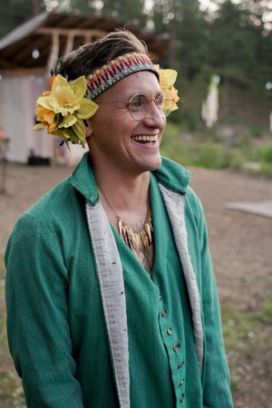 Person wearing a green jacket and floral headpiece smiles during an outdoor celebration.