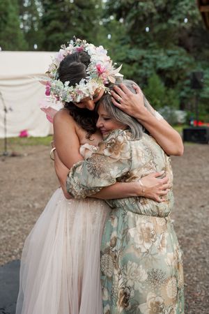 Two people share an emotional embrace while wearing floral headpieces and elegant dresses in an outdoor setting.