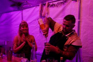 Bartenders mix drinks under dramatic purple lighting in a tent at a festive event.