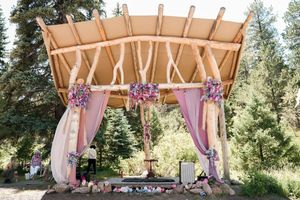 Outdoor wedding venue with wooden pergola and flowing pink curtains surrounded by tall evergreen trees.
