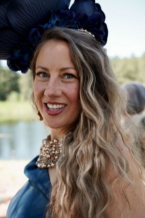 A person wearing a dark blue hat with pearls and wavy hair poses outdoors.