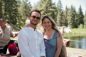 Wedding guests gather outdoors near a lake surrounded by pine trees on a sunny day.