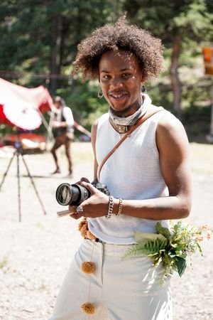 A photographer in a white outfit holding a camera and flowers at an outdoor event.