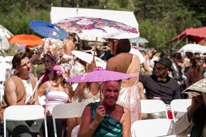 Festival-goers with parasols gather under trees during a sunny outdoor celebration.
