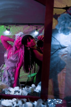 A performer in pink fairy costume and wings sings on stage with dramatic lighting effects.