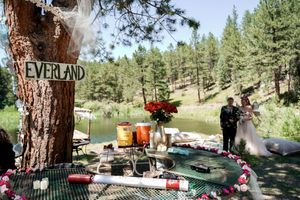 Outdoor wedding picnic setup with Neverland sign, red flowers, and green tablecloth in a pine forest setting.