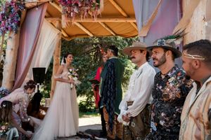 A magical outdoor wedding ceremony takes place under a wooden canopy decorated with purple flowers and draping fabric.
