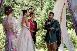Wedding ceremony taking place under an outdoor arch decorated with flowing fabric in a forest setting.