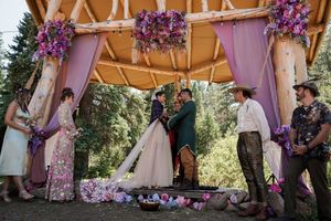 Wedding guests gather under a wooden pavilion decorated with purple flowers during an outdoor forest ceremony.