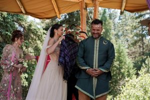 Wedding ceremony in a forest with participants in traditional and modern attire under a tan canopy.