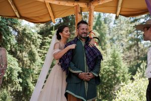 A heartfelt embrace during an outdoor wedding ceremony beneath a wooden pergola in a forest setting.