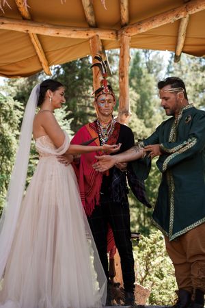 A bride in a flowing white gown participates in a traditional ceremony under a wooden canopy.