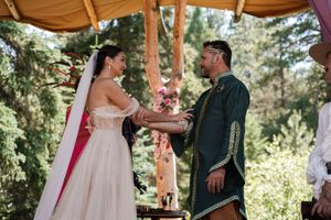 An outdoor wedding ceremony takes place under a wooden canopy surrounded by pine trees.