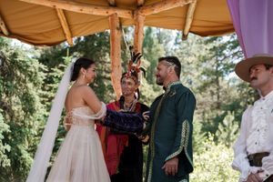 A couple shares their first kiss as newlyweds under a wooden canopy adorned with purple flowers.