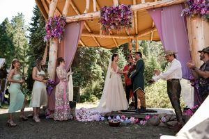 Wedding ceremony under a decorated wooden structure with purple flowers and green bridesmaids dresses.