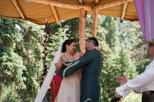A wedding ceremony takes place under a wooden canopy decorated with lights in a forest setting.