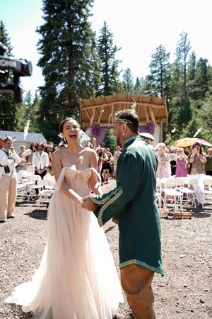 Wedding guests dance and celebrate at an outdoor forest reception with pine trees in the background.