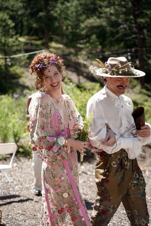A couple in whimsical wedding attire walk outdoors, with one in a floral dress and another in vintage-style clothing.