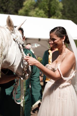 A bride in a white dress pets a white unicorn-styled horse during an outdoor celebration.