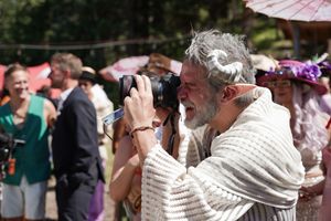 A photographer with a white scarf and grey beard photographs an event outdoors among a crowd.