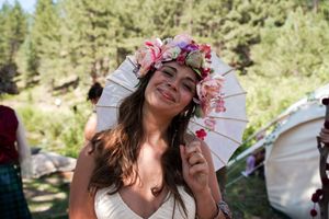 Bride wearing floral crown and holding parasol smiles joyfully during outdoor forest wedding.