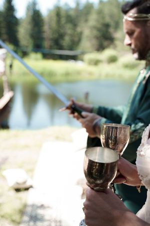 Close-up of wine glasses and hands holding drinks by a serene lake with pine trees in background.