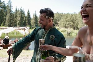Two people laughing while holding drinks at an outdoor celebration in a forest setting.