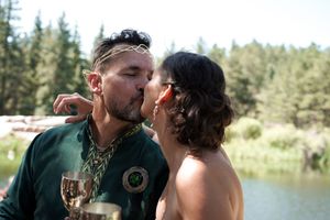 A couple sharing a playful moment with wine glasses by a serene lake.