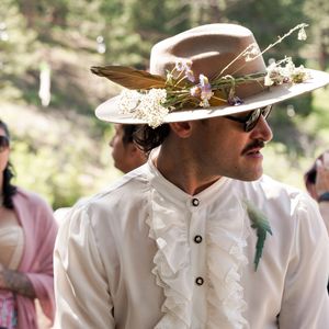 Person wearing ornate white ruffled shirt and decorated wide-brimmed hat at outdoor gathering.