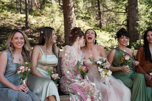 Wedding party members seated outdoors holding bouquets and wearing various colored dresses.