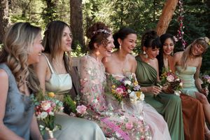 Wedding party members gather outdoors in pastel and floral dresses under forest trees.