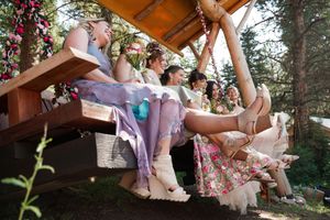 Wedding party sits casually on wooden deck, swinging their feet and laughing together in forest setting.