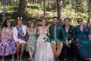 A wedding party sits together on a rustic wooden bench in a forest setting wearing colorful formal attire.