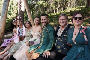 Wedding guests smile and relax on a wooden bench decorated with pink flowers in an outdoor forest setting.