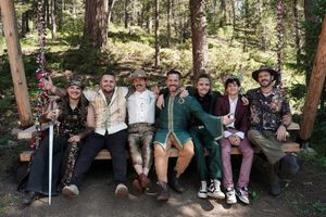 A group photo of friends in eclectic outfits sitting together on a wooden bench in a forest setting.
