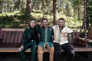 Three people sit together on a wooden bench in a forest setting during what appears to be a festive outdoor gathering.