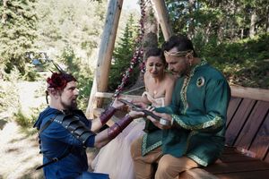 Three people in festive attire share a moment on wooden bench during outdoor wedding celebration.